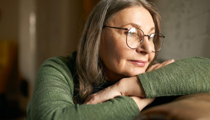 Close up image of beautiful fashionable mature woman wearing stylish round eyeglasses relaxing indoors on leather couch, having pensive deep in thoughts facial expressions, thinking about her life